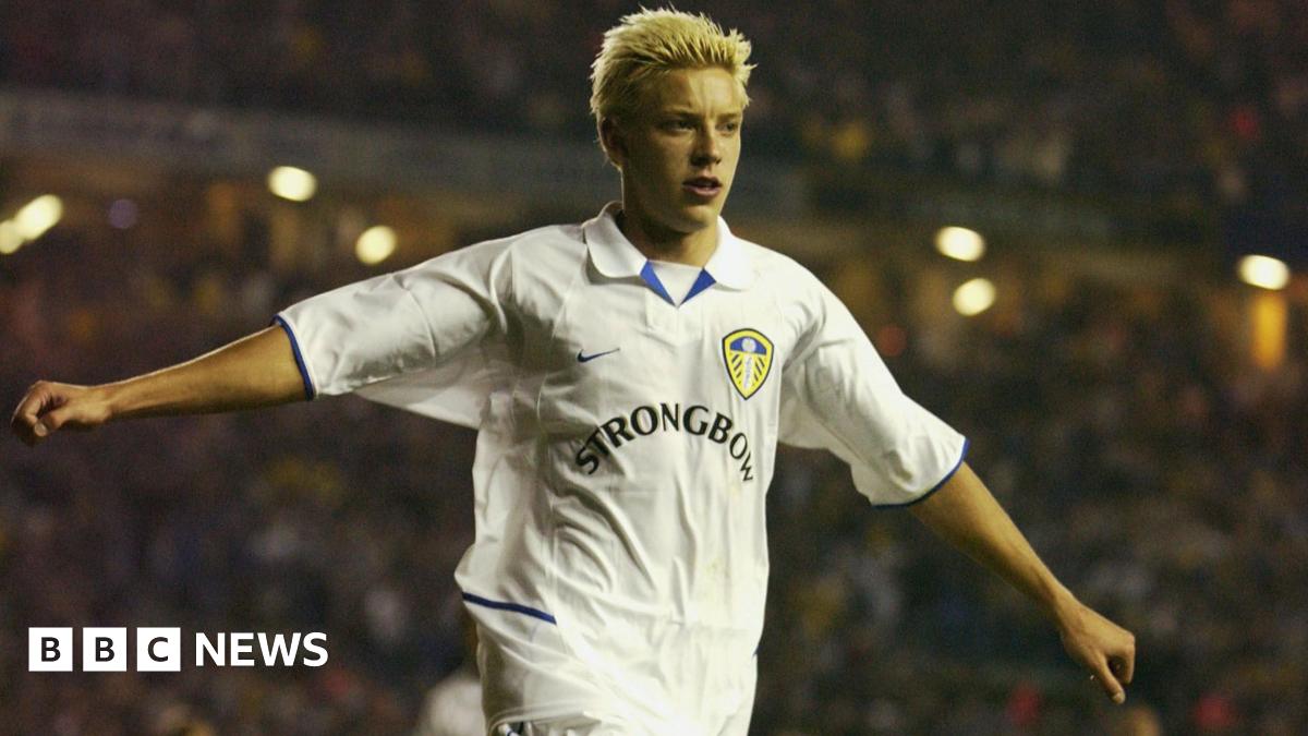 A young Alan Smith celebrating a goal for Leeds United, with his arms outstretched. He has spiky blond hair and the 'Strongbow' logo is prominent on the jersey.