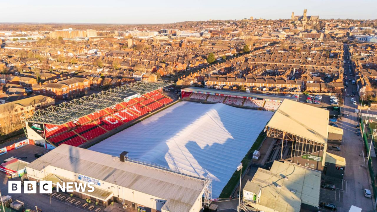 Aerial view of a football stadium with a protective cover, resembling a huge white tent, over the playing surface. There are four stands surrounding the pitch. The word "city" can be seen on one of the stands, which has red seats, and there are houses and a large cathedral in the background.