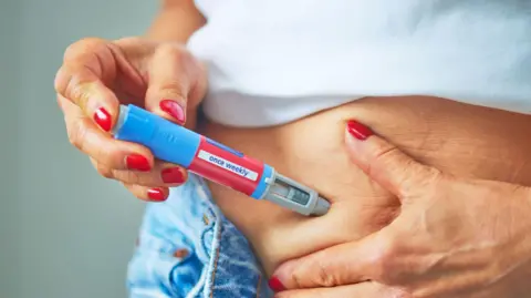 Getty Images A woman, wearing bright red nail polish and unbuttoned blue jeans, injects herself into the skin and soft tissue of her lower abdomen with an obesity jab pen. 