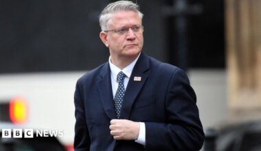 Close up of Andrew Rosindell walking outside wearing a suit with a union jack flag pin on the lapel . He is looking to his left