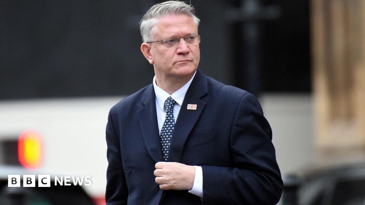Close up of Andrew Rosindell walking outside wearing a suit with a union jack flag pin on the lapel . He is looking to his left