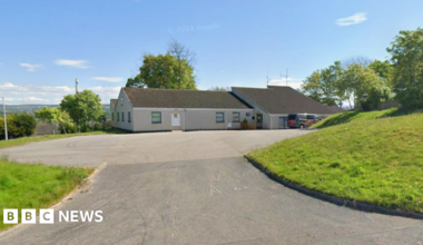 A grey building in Inverness with a black roof in the background with green grass to the right and left, in front of a bright blue sky.