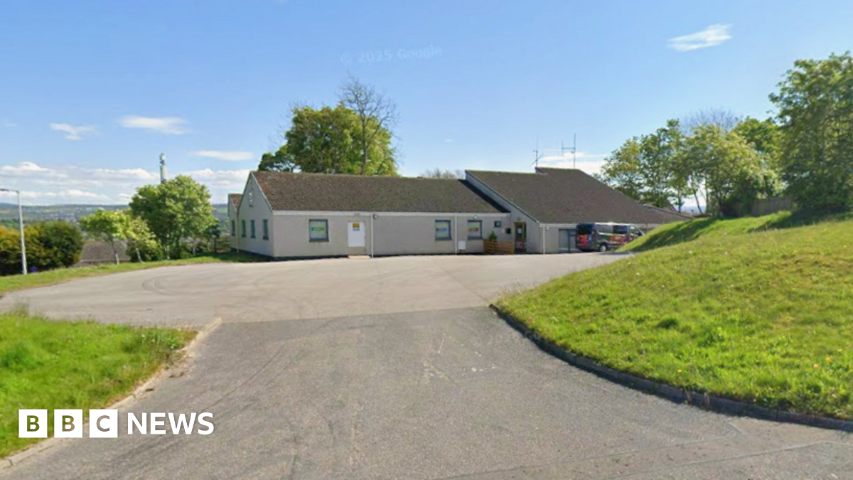 A grey building in Inverness with a black roof in the background with green grass to the right and left, in front of a bright blue sky.