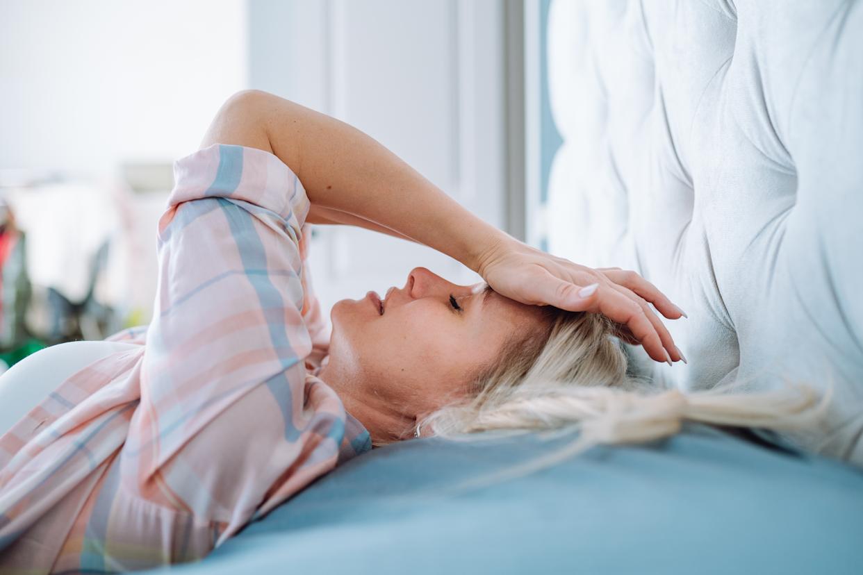 Struggling blonde woman with a severe headache, lying on bed in light bedroom.