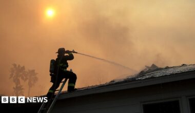 Millie Bobby Brown and David Harbour in character as Eleven and Jim Hopper. Eleven wears red shorts over grey leggings and a grey jumper. Jim Hopper wears a black leather jacket, a black beanie hat and dark trousers.They are speaking to director Frank Darabont on the set of Stranger Things. There is a vintage advertisement posted in the background with debrief on the ground.