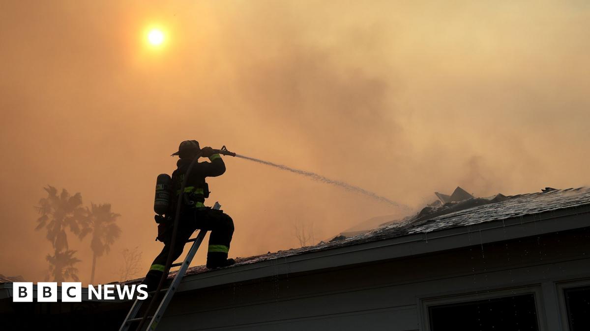 Millie Bobby Brown and David Harbour in character as Eleven and Jim Hopper. Eleven wears red shorts over grey leggings and a grey jumper. Jim Hopper wears a black leather jacket, a black beanie hat and dark trousers.They are speaking to director Frank Darabont on the set of Stranger Things. There is a vintage advertisement posted in the background with debrief on the ground.