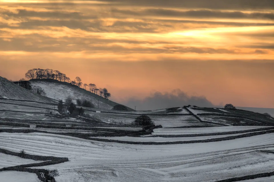 Sunset on a winter evening in the Yorkshire Dales National park with snowy fields bounded by stone walls in the foreground backed by orange sunlight through broken clouds.