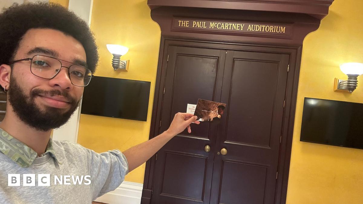 A male student at LIPA in Liverpool holding the postcard in front of a door which reads 'the paul mccartney auditorium'