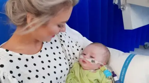 Ann Reel Hollie as a newborn baby in her mum Ann's arms. She has tubes in her nose and tubes around her body. She has a green babygrow on. Ann has her blonde hair tied back and is wearing a black and white polka dot top. 