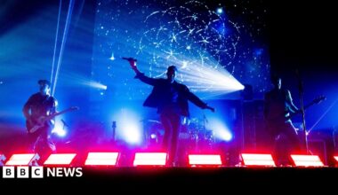 Three members of Coldplace performing on stage/ Two are holding guitars and the man in the middle is holding a microphone. The stage lights are blue and red.