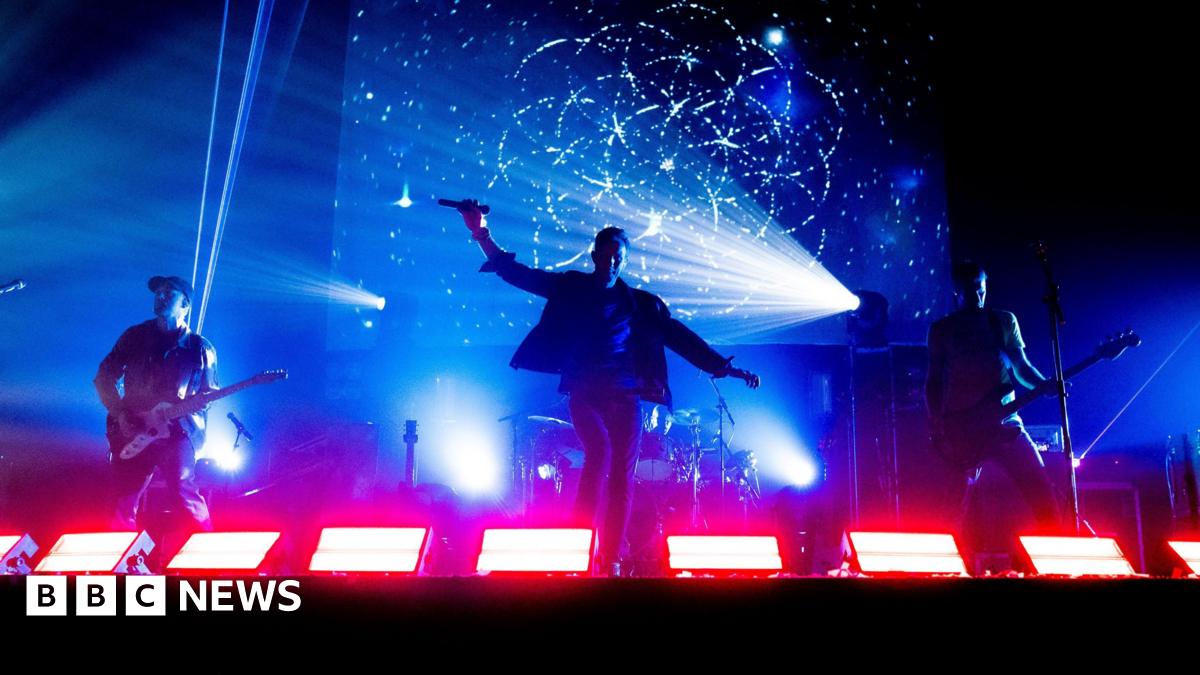 Three members of Coldplace performing on stage/ Two are holding guitars and the man in the middle is holding a microphone. The stage lights are blue and red.