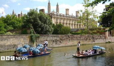 Punting in Cambridge. Three punts can be seen next to a walled section of the river. A university college building is in the background, and the spires or King's College Chapel.