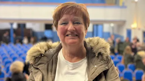 A woman with light brown hair, wearing a white top and grey jacket, looks at the camera