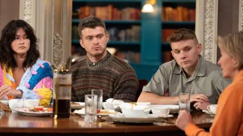 BBC/Studio Lambert/Euan Cherry Two men sit at a table with food plates and cafetiere in front of them, with two women on either side. The three on the left look towards the woman on the right, who is seen in profile, speaking. She is wearing an orange jumper and has short blonde hair. The woman on the far left has shoulder length dark hair and is wearing a blue, pink, and yellow chunky knit cardigan