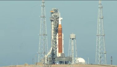 An orange and white rocket is held vertically by scaffolding on a launch pad.