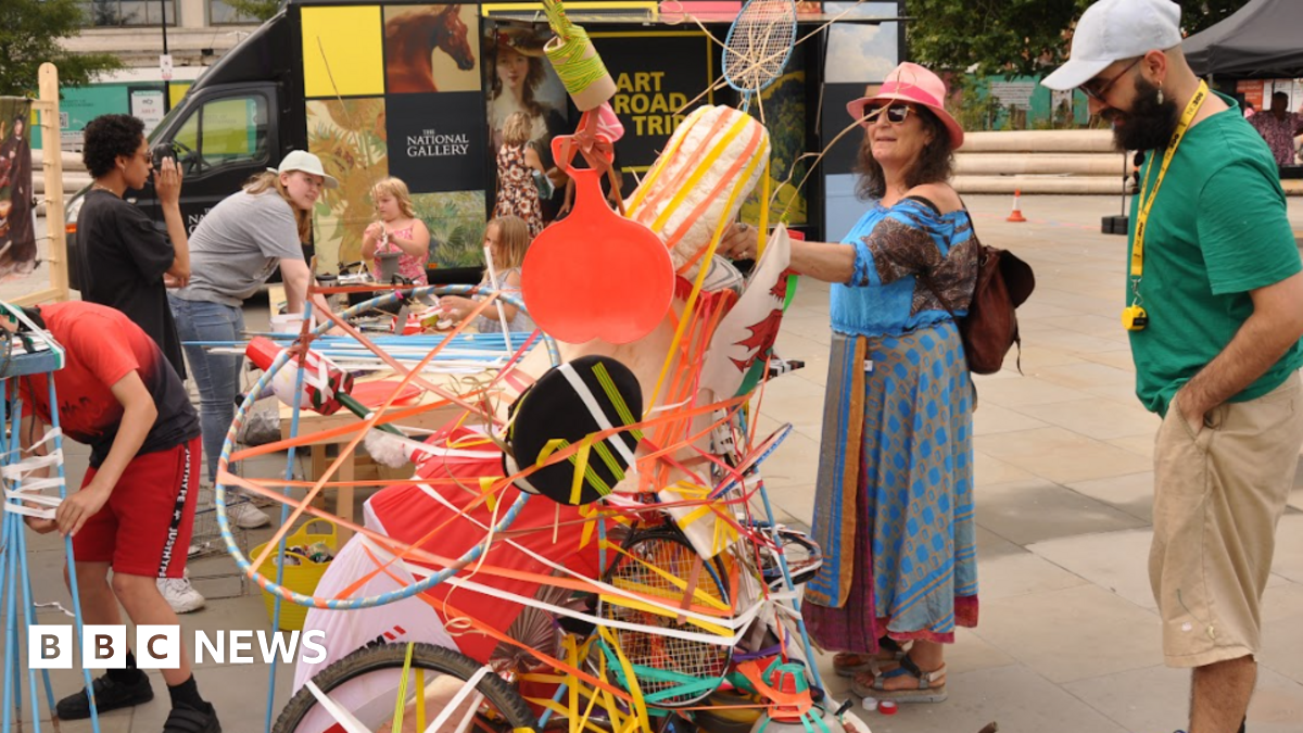 A quirky art piece made up of colourful objects such as tennis rackets and hoola hoops, pieced together. Two people are admiring the piece, a woman in a long blue dress with sunglasses and a pink hat and a man in a green top and beige shorts with a grey baseball cap. The art piece is outside, other activities are taking place in the background.