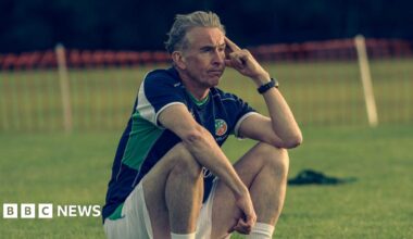 Steve Coogan sitting on the sidelines in a Republic of Ireland training kit looking disconsolate. He has grey hair and has his left elbow leaning on his knee.