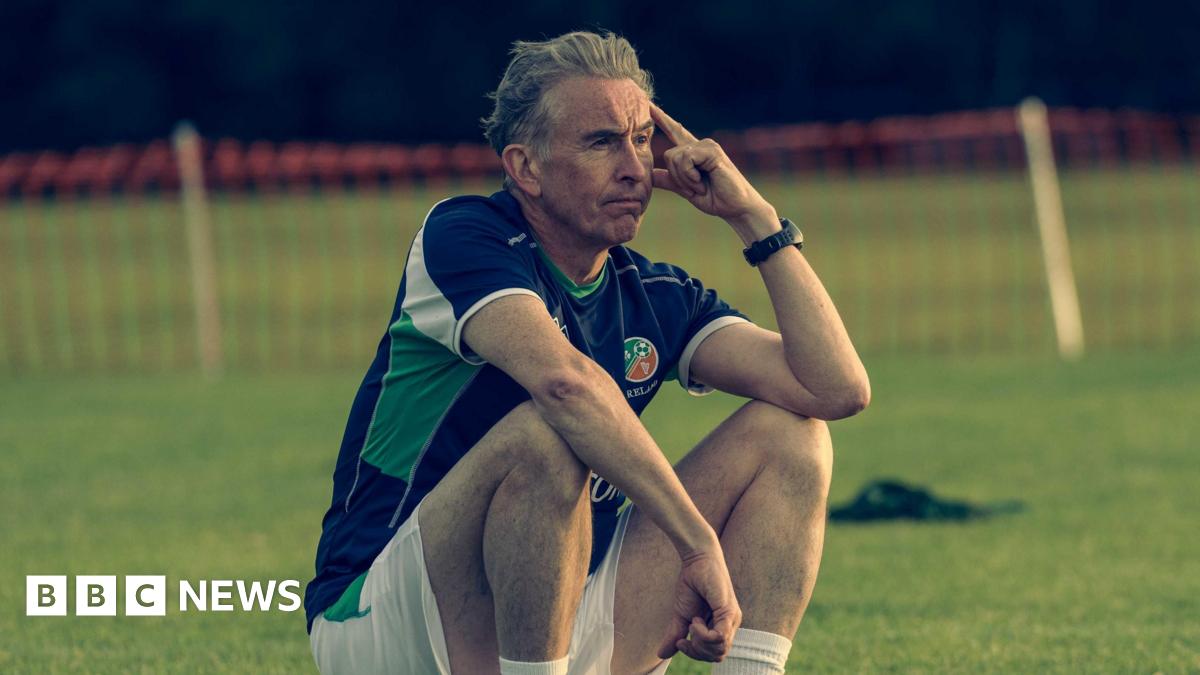 Steve Coogan sitting on the sidelines in a Republic of Ireland training kit looking disconsolate. He has grey hair and has his left elbow leaning on his knee.