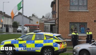 Two uniformed members of the Gardaí standing nearby a property in the Cherry Orchard area of Ballyfermot where the body of a man in his 40s was found.  There is a beige-brick house in the foreground and a row of white semi-detached houses in the background.  A yellow and blue Garda car is parked at the scene along with other vehicles. Irish tricolours fly from two lamp posts.