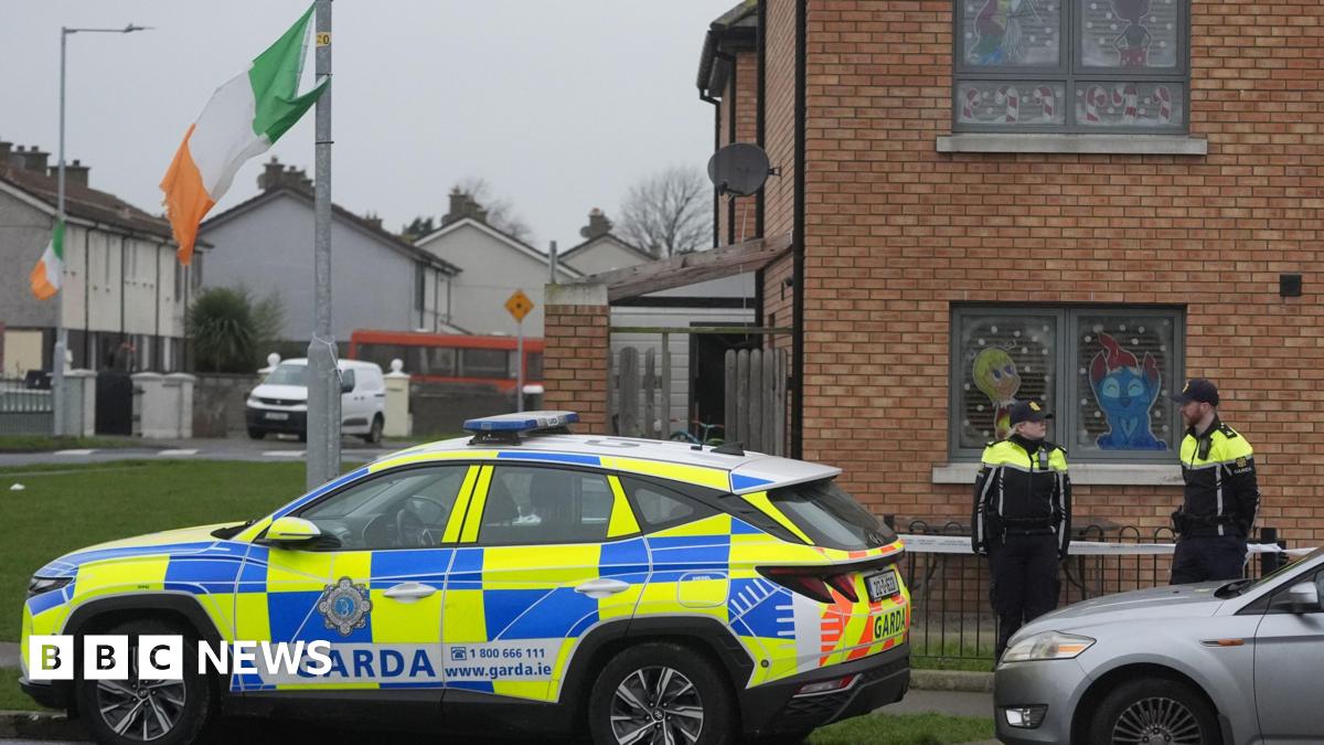 Two uniformed members of the Gardaí standing nearby a property in the Cherry Orchard area of Ballyfermot where the body of a man in his 40s was found.  There is a beige-brick house in the foreground and a row of white semi-detached houses in the background.  A yellow and blue Garda car is parked at the scene along with other vehicles. Irish tricolours fly from two lamp posts.