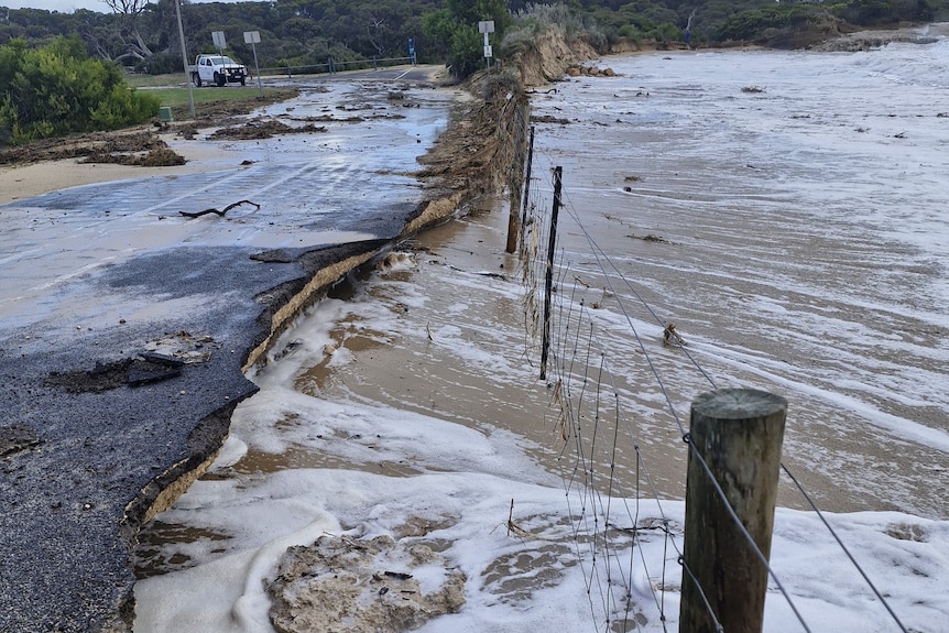 A water damaged road with waves coming under a fence and lapping against it.