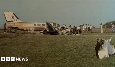 An old photograph shows the wreckage of a small aircraft in a grassy field while people dig through its remains and others watch on.