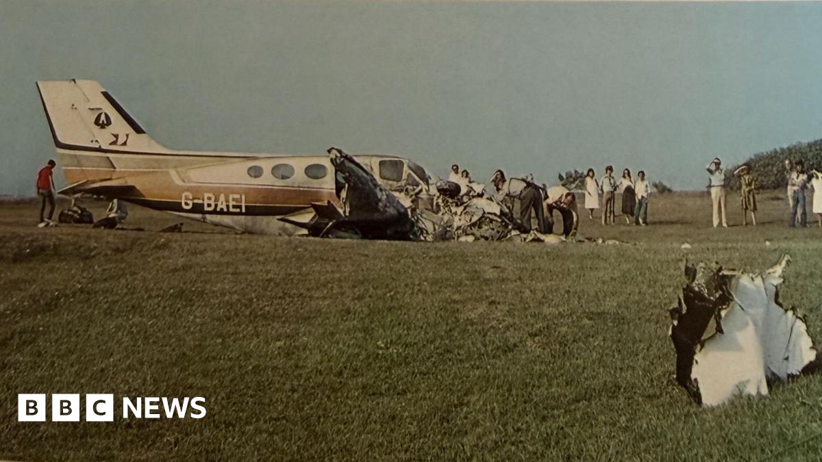 An old photograph shows the wreckage of a small aircraft in a grassy field while people dig through its remains and others watch on.
