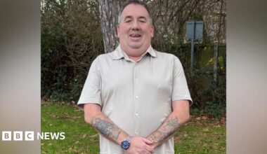 A man wearing a beige shirt is standing in a garden. He has tattoos all over his arms and is wearing a watch.