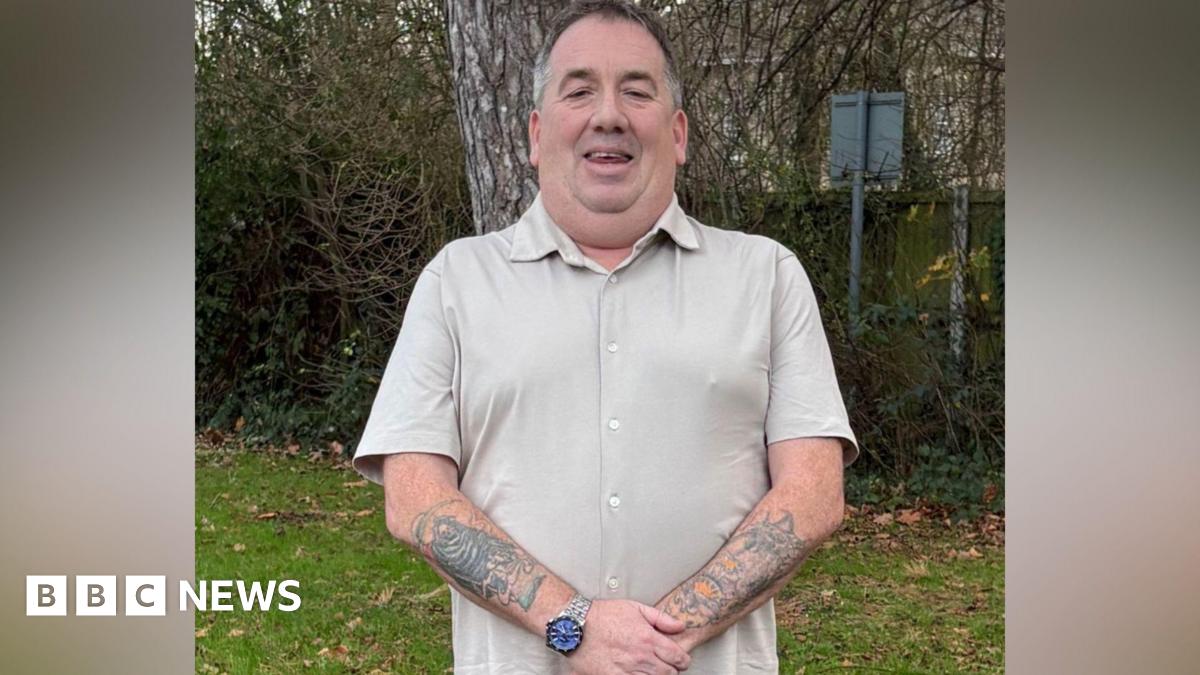 A man wearing a beige shirt is standing in a garden. He has tattoos all over his arms and is wearing a watch.