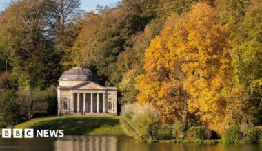 Stourhead in Wiltshire. There are orange, green and yellow trees in the shot and a building overlooking water.