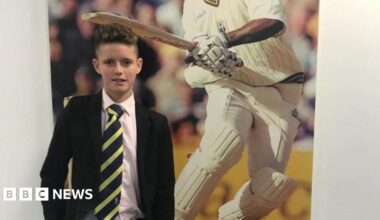 A schoolboy in a black blazer, white shirt and navy and yellow diagonally striped tie standing in front of a poster showing a cricketer.