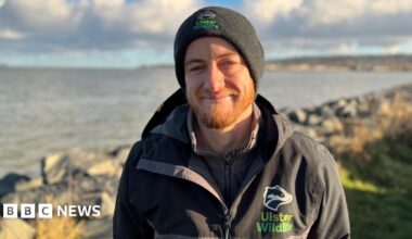 A man standing in front of Belfast Lough. He is wearing a black and grey rain jacket with an Ulster Wildlife logo on it. He is also wearing a black hat.