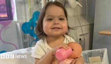 A little girl in a hospital room smiles as she feeds a doll from a bottle.