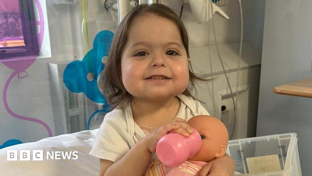 A little girl in a hospital room smiles as she feeds a doll from a bottle.