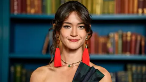 Studio Lambert/BBC/PA A head and shoulders photo of a young woman with dark hair tied back, with loose curls around her face. She is wearing red earrings with long tassels, a green jewelled choker necklace and the top of a tartan bow on a shoulderless corset can just be seen. Behind her are blue painted shelves with books.