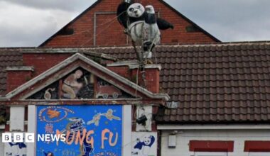 An ornamental figure of a black and white panda kicking one leg in the air. The figure is on the roof of a building which has a blue sign on the wall which says 'Kung Fu'.