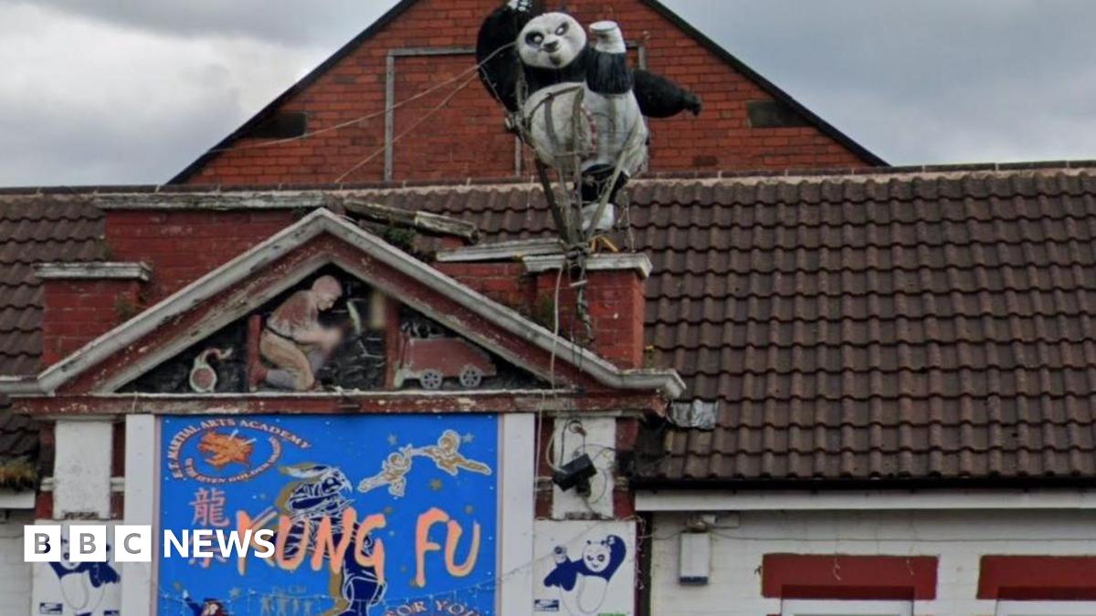 An ornamental figure of a black and white panda kicking one leg in the air. The figure is on the roof of a building which has a blue sign on the wall which says 'Kung Fu'.