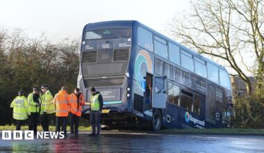 The Stagecoach bus is across a grass verge with its front wheels in a ditch and trees and houses in the background. Police are at the scene in hi-vis jackets and there are more people in orange jackets. The ground is wet.