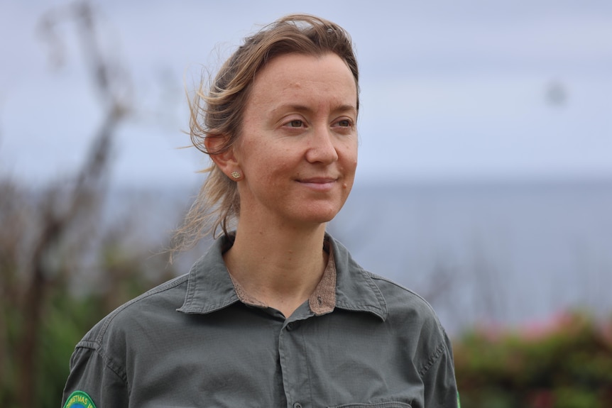 A woman smiles looking off into the middle distance, the ocean and an overcast sky behind her.