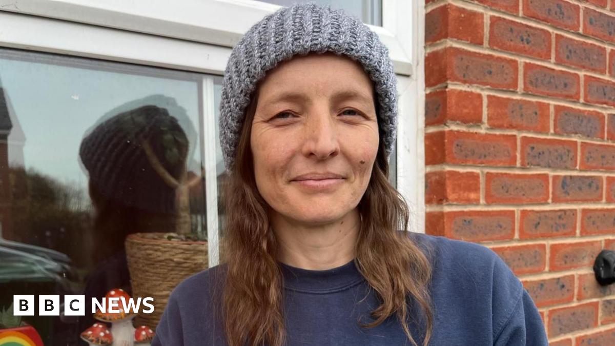 Lucy in a grey hat. She has brown hair and is also wearing a blue jumper. She is stood in front of a window.