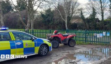 A marked blue and yellow Essex Police car is parked on a muddy road. A red and black quad bike is parked in front of it. Behind the bike are green metal railings that look like they lead to a park.