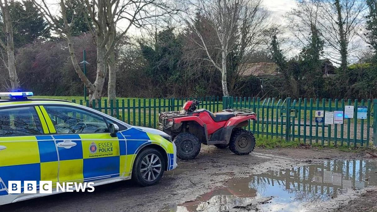 A marked blue and yellow Essex Police car is parked on a muddy road. A red and black quad bike is parked in front of it. Behind the bike are green metal railings that look like they lead to a park.