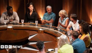 Contestants on The Traitors sitting behind a round table during an episode. One woman can be seen talking as the others listen to her.