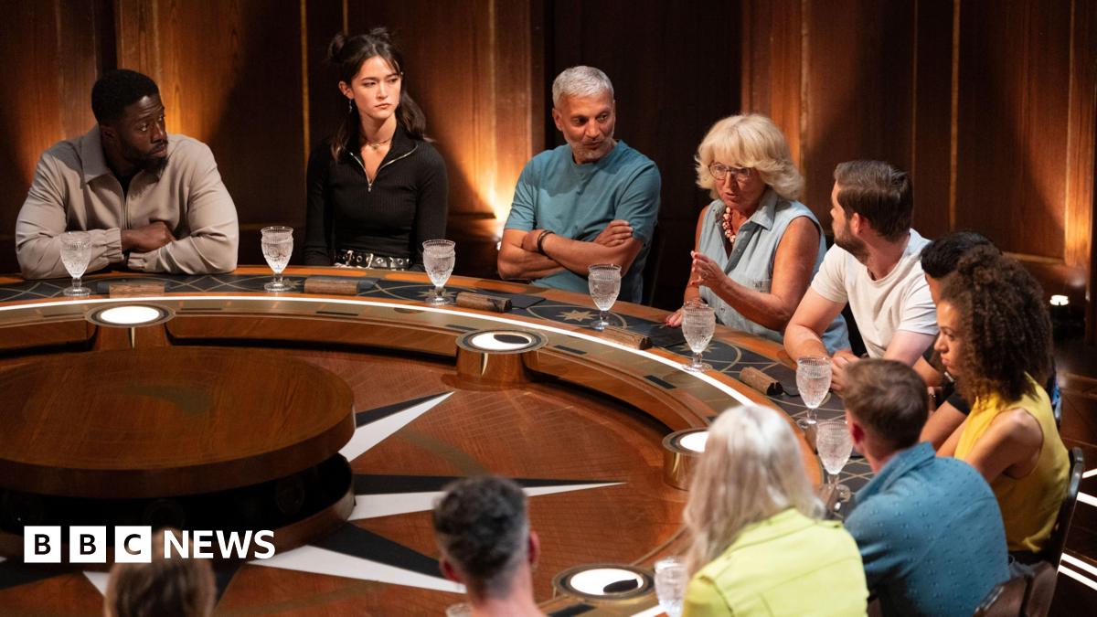 Contestants on The Traitors sitting behind a round table during an episode. One woman can be seen talking as the others listen to her.