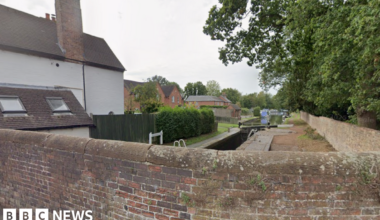 A view from a brick bridge which is built over a canal. The view shoes the canal stretching off into the distance with trees to the right of it and red brick houses to the left.