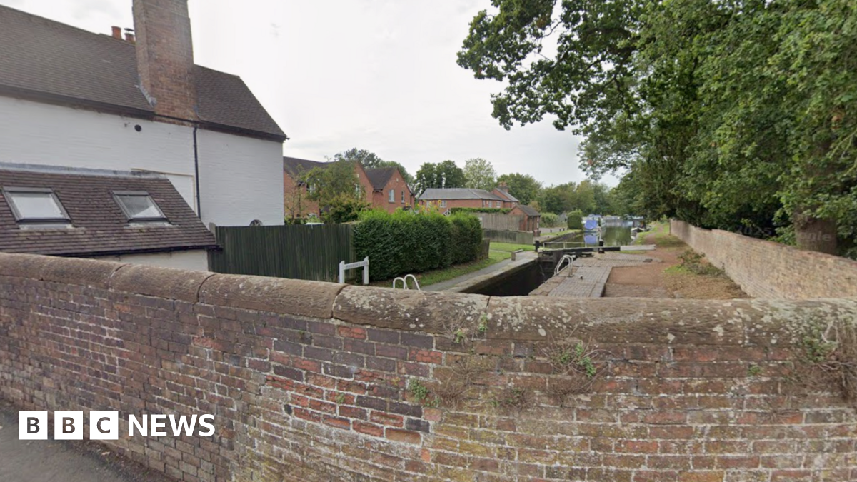 A view from a brick bridge which is built over a canal. The view shoes the canal stretching off into the distance with trees to the right of it and red brick houses to the left.