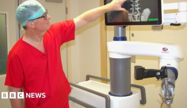 Dr Simon Tizzard is wearing a blue medical hat, orange scrubs and glasses pointing at a spine X-ray on the screen of the robot in a hospital room.