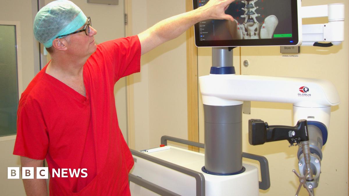 Dr Simon Tizzard is wearing a blue medical hat, orange scrubs and glasses pointing at a spine X-ray on the screen of the robot in a hospital room.