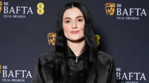 Reuters Posy Sterling with long black hair and a black jacket, in front of a Bafta-branded backdrop
