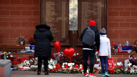 Reuters People stand in front of the Hillsborough Memorial outside Anfield Stadium in Liverpool. There are flowers and heart balloons in front of a plaque with the names of the 96 victims of the disaster.